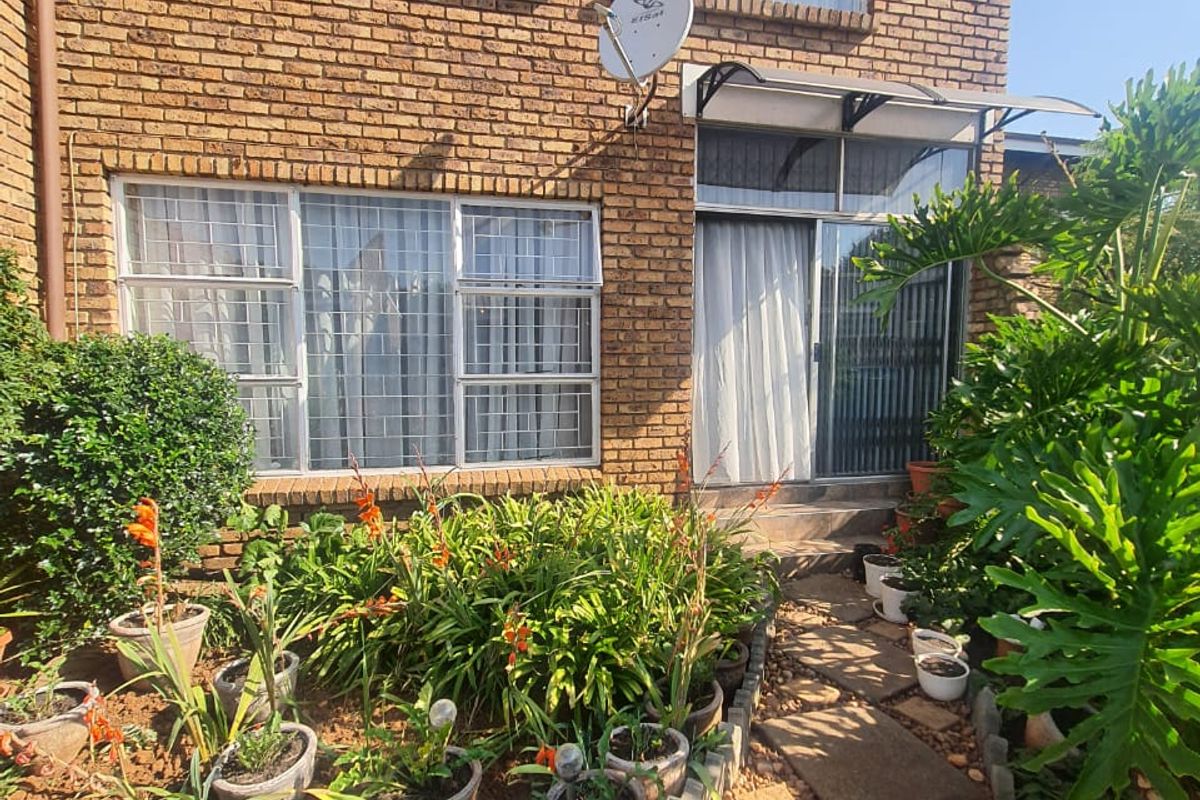 Rear view of a double-storey face brick townhouse featuring a paved walkway, established garden beds, and secure aluminium window frames.