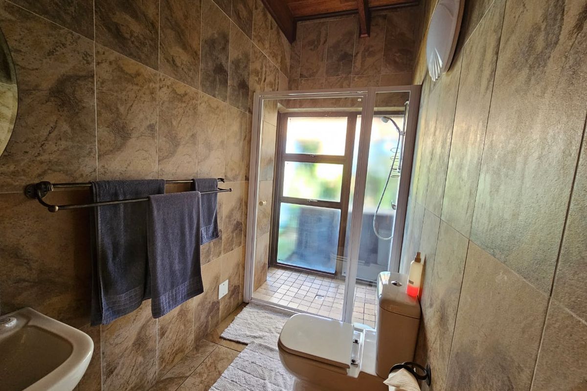 A compact bathroom featuring full-height wall tiling, a shower enclosure, toilet, washbasin, and exposed timber ceiling beams, providing a rustic aesthetic.