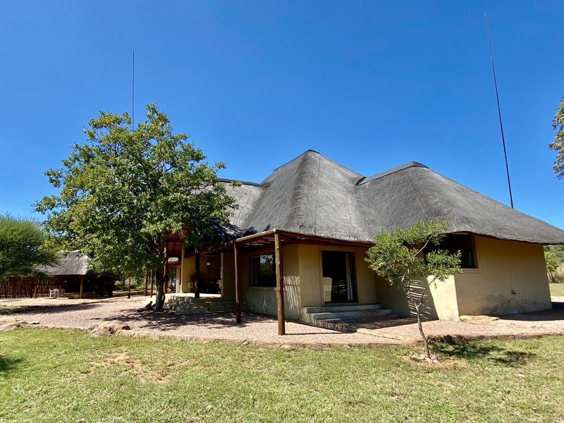 Rear view of a traditional thatch-roofed house featuring a covered veranda and gravel patio area set within a natural garden landscape.
