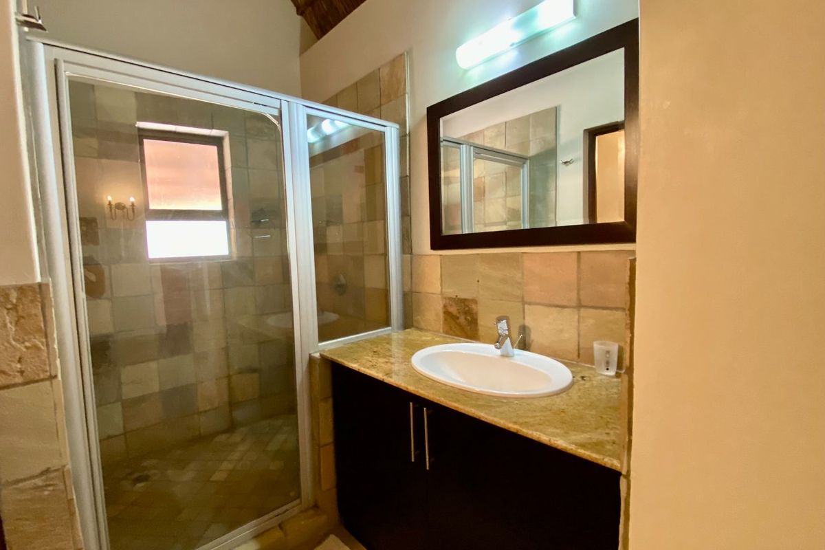 A bathroom featuring a shower enclosure, a vanity cabinet with a stone countertop, a washbasin, and a mirror under a high thatched ceiling.