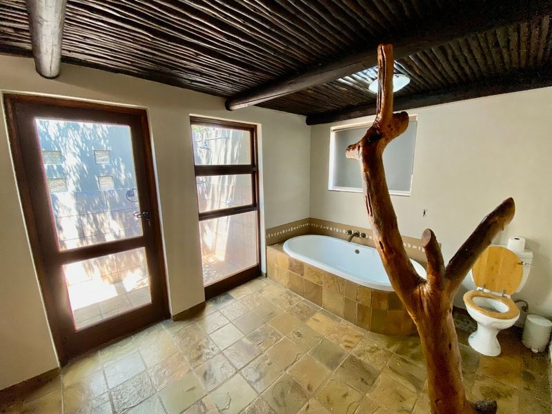 A rustic-style bathroom featuring a built-in bathtub with stone-tiled surround, exposed timber beam ceiling, and direct access to an outdoor shower area.