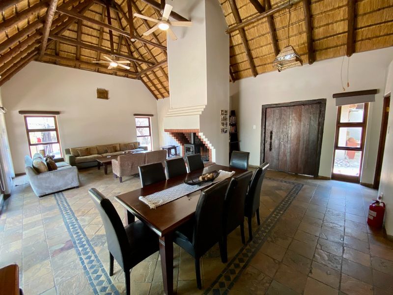 Spacious open-plan living and dining area featuring a high vaulted thatched ceiling, exposed timber beams, stone tile flooring, and a central brick fireplace.