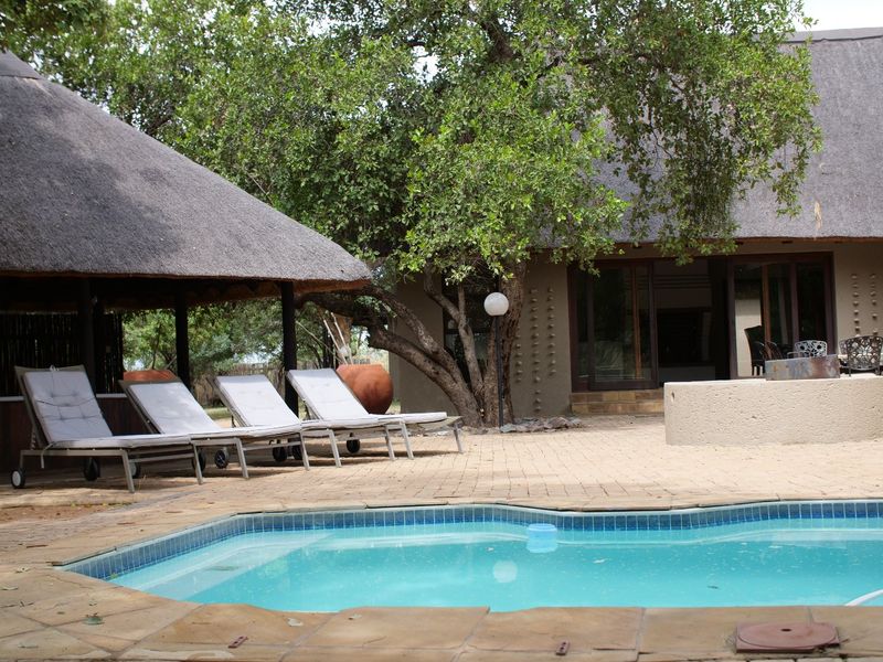 Outdoor pool area featuring a paved deck, thatched lapa, and established trees, providing a relaxed setting for outdoor leisure.