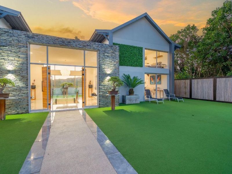 Modern rear exterior view featuring stone cladding, expansive glass doors, a well-maintained artificial lawn, and a concrete pathway leading to the home.
