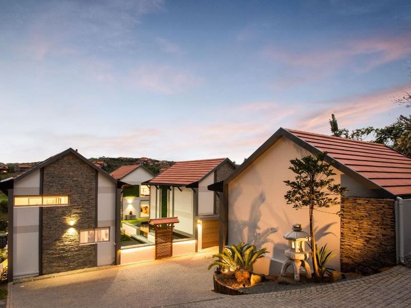 A modern residential property featuring stone-clad facades, tiled roofs, and a paved driveway, captured during twilight with integrated exterior lighting.