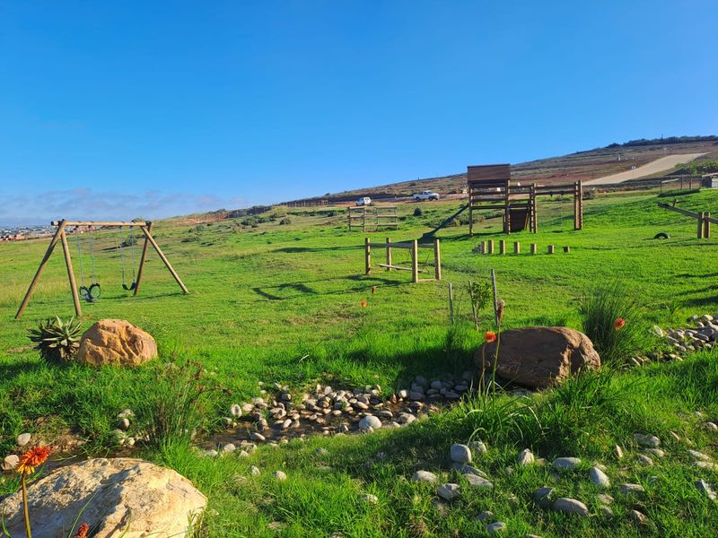Expansive green garden area featuring a children's playground, natural rock landscaping, and a small drainage channel set against a scenic hillside backdrop.