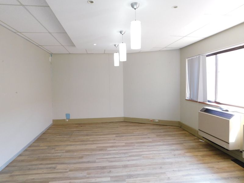 A bright, empty office space featuring wood-look laminate flooring, a suspended ceiling with integrated lighting, and a wall-mounted HVAC unit.