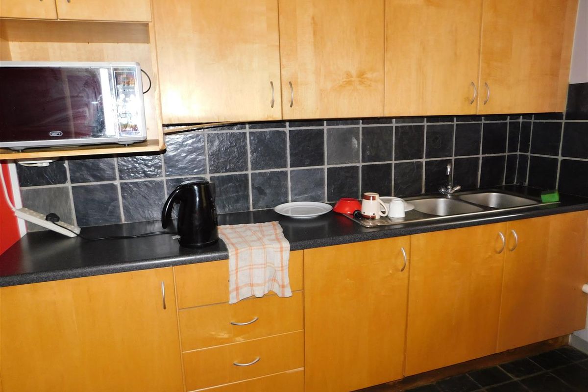 A functional office kitchenette featuring light wood cabinetry, dark stone-look countertops, and a black tiled backsplash with a double stainless steel sink.