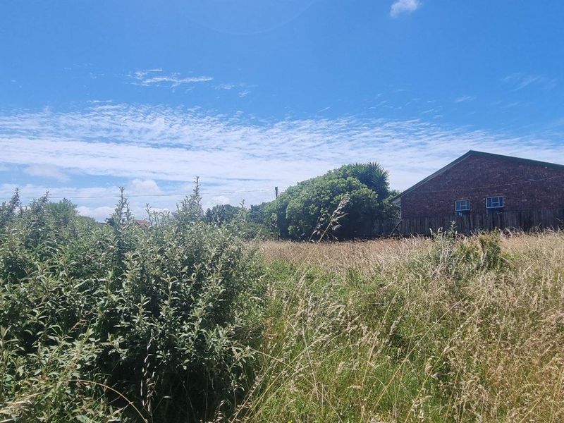 A vacant residential plot featuring overgrown grass and shrubs, bordered by existing residential fencing and neighboring brick structures.