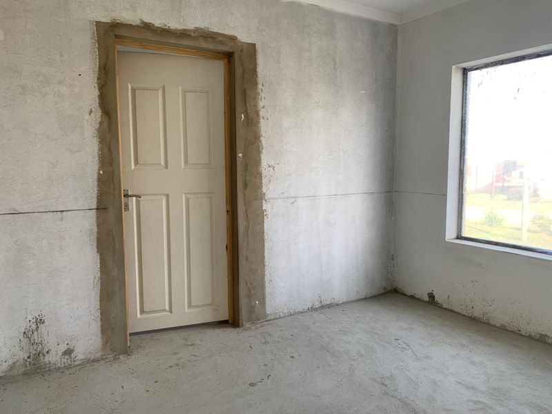 An unfinished bedroom interior featuring raw plaster walls, an installed panel door, and a window, currently in a shell condition awaiting final finishes.
