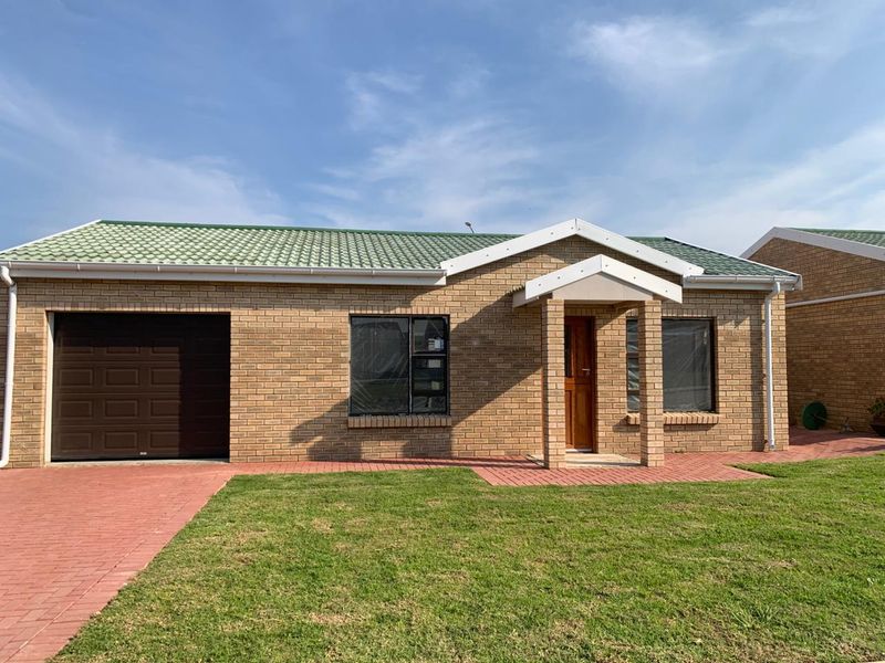 A well-maintained single-story face brick retirement unit featuring a green tiled roof, single garage, and a paved entrance path.