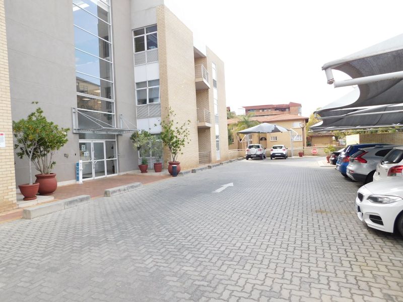 Exterior view of a modern office building featuring a paved parking area with covered bays and a prominent glass entrance facade.