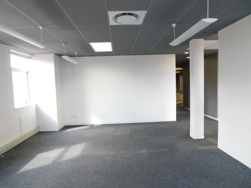 A bright, vacant office space featuring a suspended ceiling, recessed lighting, carpeted flooring, and a central structural column.