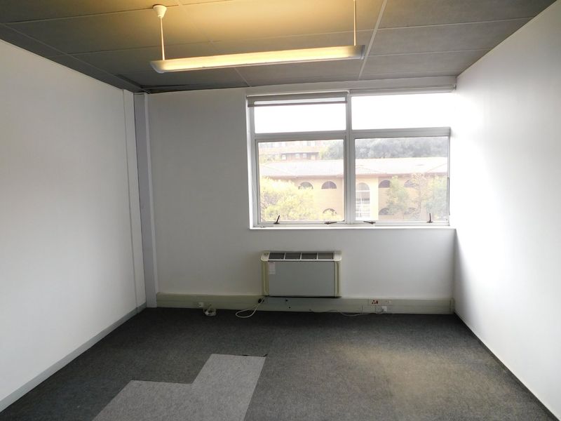 A vacant office space featuring a suspended ceiling, carpeted flooring, and a wall-mounted air conditioning unit with natural light from a large window.