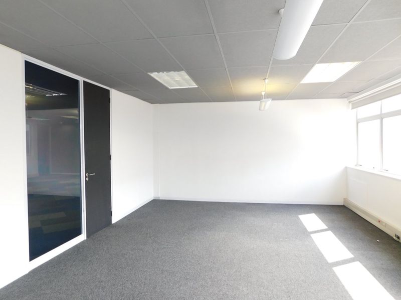 A bright, vacant office space featuring a suspended ceiling, recessed lighting, carpeted flooring, and a modern glass partition door.