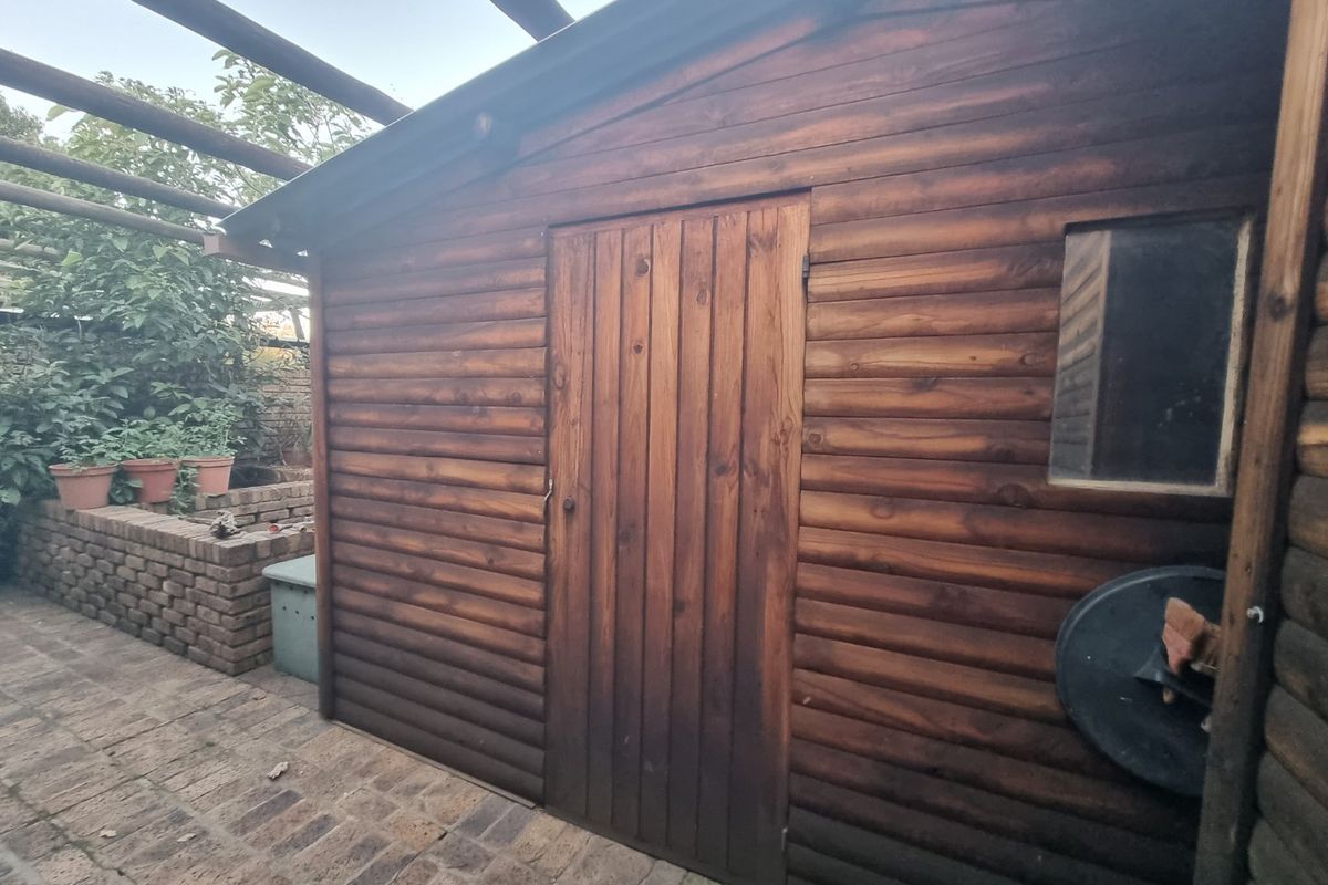 A timber garden shed featuring a wooden door and window, situated on a paved patio area with a brick planter.