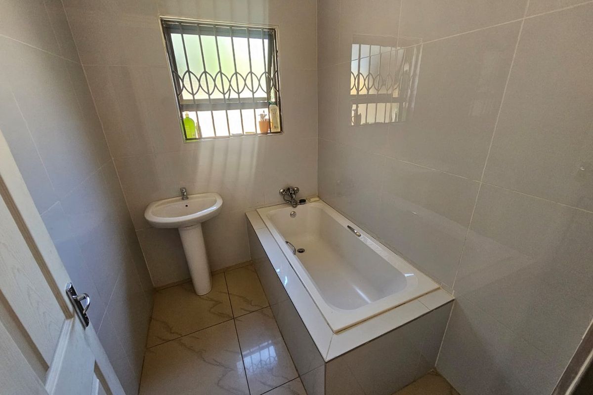 A functional bathroom featuring a built-in bathtub, pedestal basin, and floor-to-ceiling wall tiling with a security-grilled window.