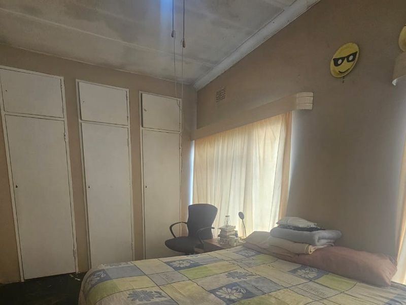 A bedroom featuring built-in cupboards and large windows with curtains, showing signs of wear on the ceiling and walls.