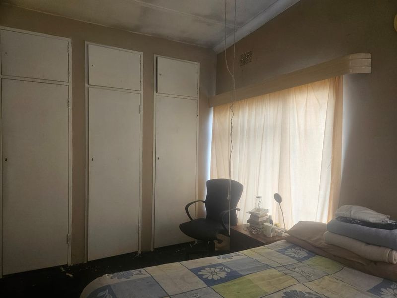 A bedroom featuring built-in cupboards and a window with curtaining, showing signs of wear on the ceiling and flooring.