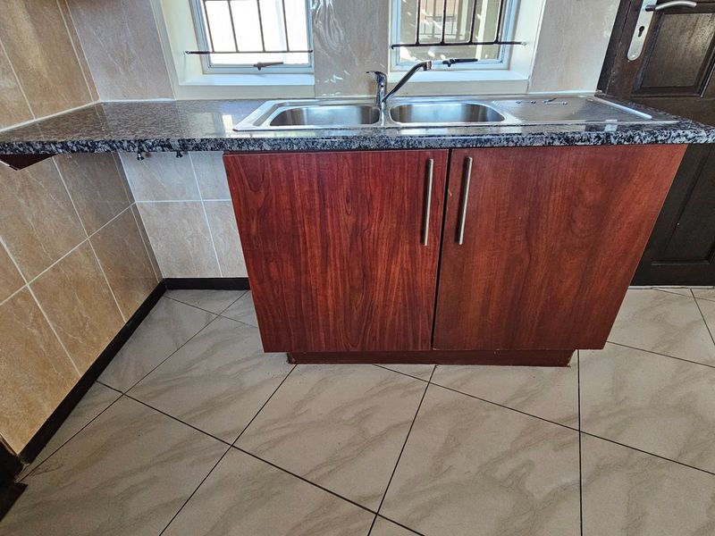 A functional kitchen area featuring a double stainless steel sink, granite countertop, and wood-finish cabinetry with tiled flooring and wall splashbacks.