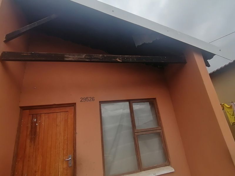 Front facade of the property showing a timber entrance door and window, with significant damage to the roof structure and missing roof sheeting.