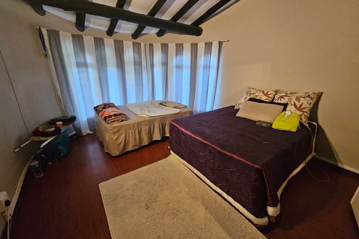 A bedroom featuring exposed timber ceiling beams and large windows with curtains, providing a rustic aesthetic with wood-look flooring.