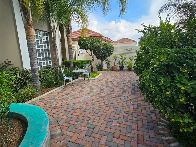 A well-maintained paved patio area featuring established garden beds, mature trees, and a decorative curved planter wall.
