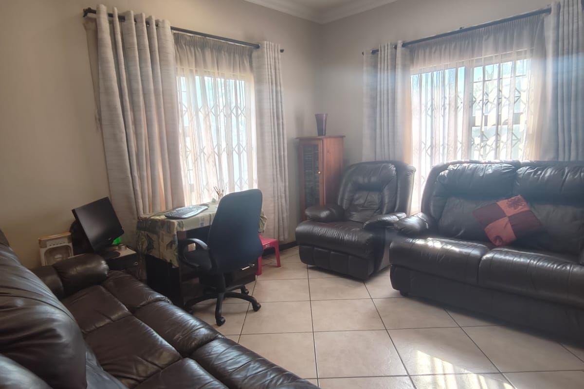 A spacious living room featuring neutral tiled flooring, recessed downlighting, and large windows fitted with security gates and curtains.