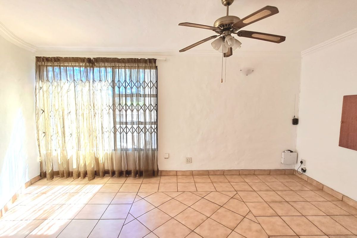 A spacious living area featuring tiled flooring, a ceiling fan, and a large window with security gate, offering a bright and functional living space.