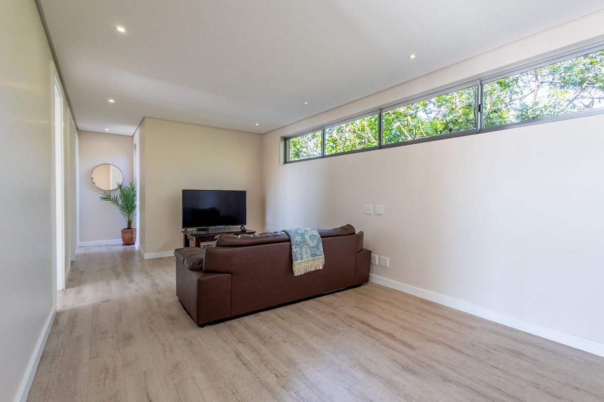 A bright living room featuring modern wood-look flooring, recessed ceiling lighting, and a high-set horizontal window providing natural light and garden views.