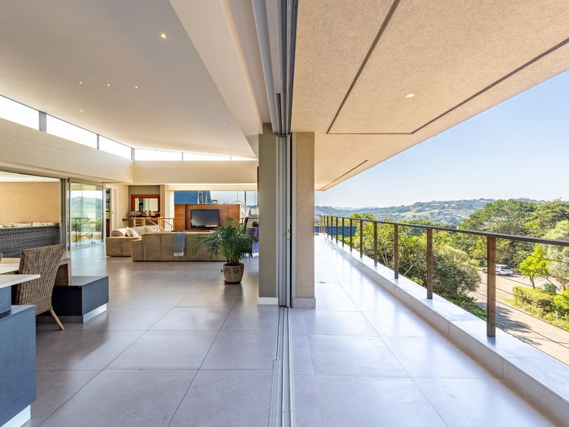 Expansive open-plan living area seamlessly flowing onto a large covered balcony with glass balustrades and panoramic hillside views.