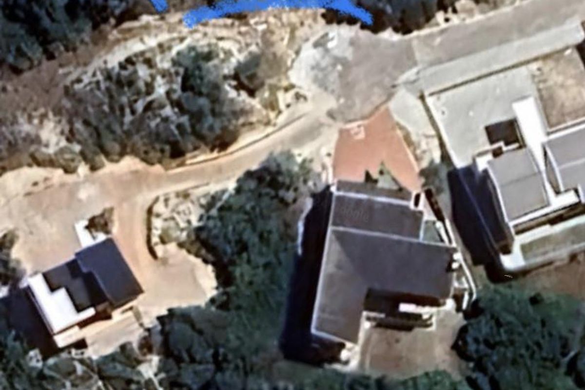 Aerial view of a vacant residential plot outlined in blue, situated within a developed neighborhood with surrounding vegetation and nearby existing properties.