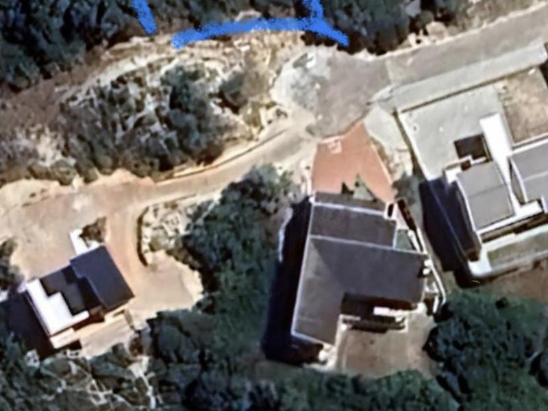 Aerial view of a vacant residential plot outlined in blue, situated within a developed neighborhood with surrounding vegetation and nearby existing properties.