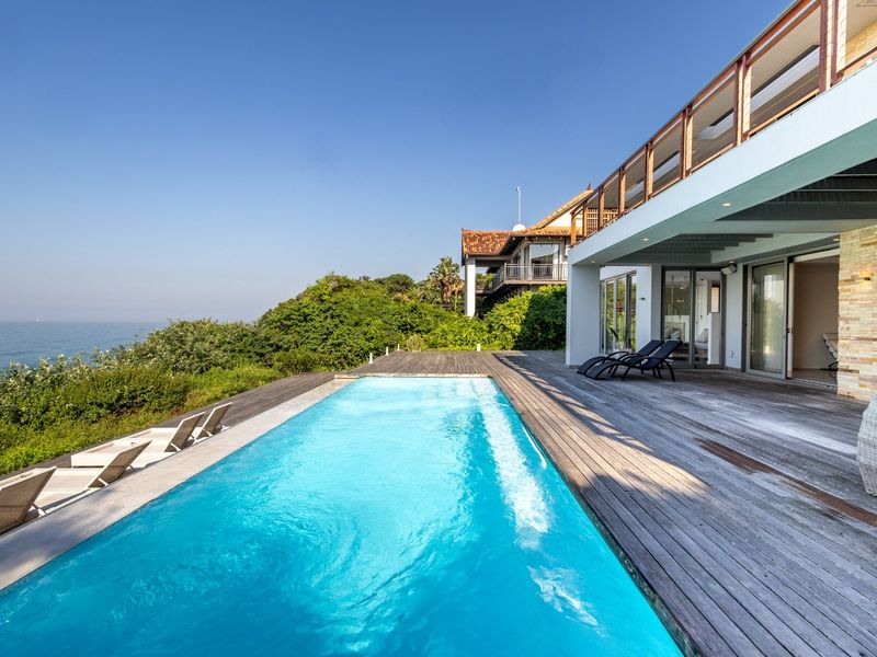 A stunning outdoor pool area featuring extensive timber decking, stone accents, and direct access to the home, overlooking a scenic coastal landscape.