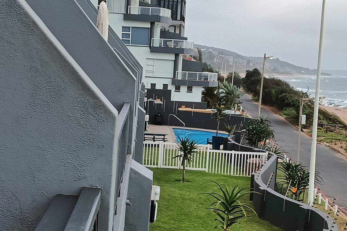 An elevated view of the apartment complex exterior, featuring a swimming pool, landscaped garden area, and secure perimeter fencing near the coastline.