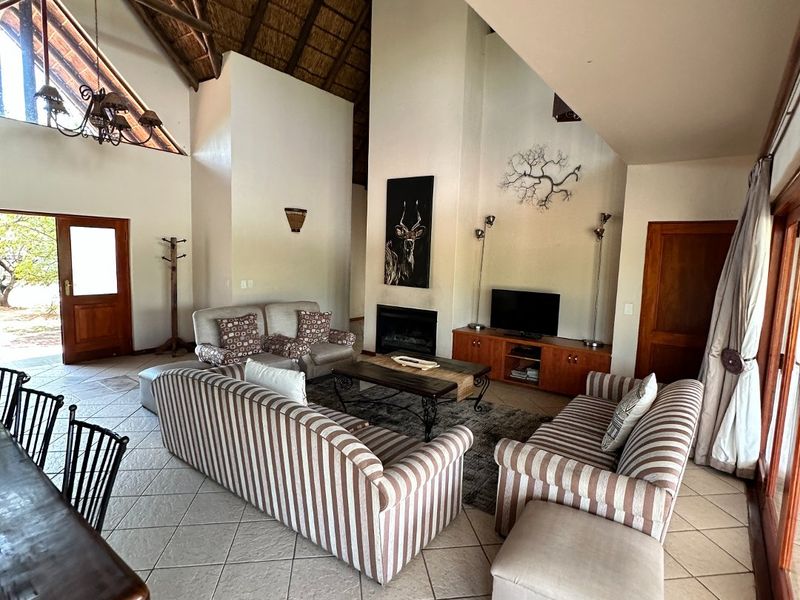 Spacious open-plan living area featuring a high vaulted thatched ceiling with exposed timber trusses, tiled flooring, and a prominent built-in fireplace.