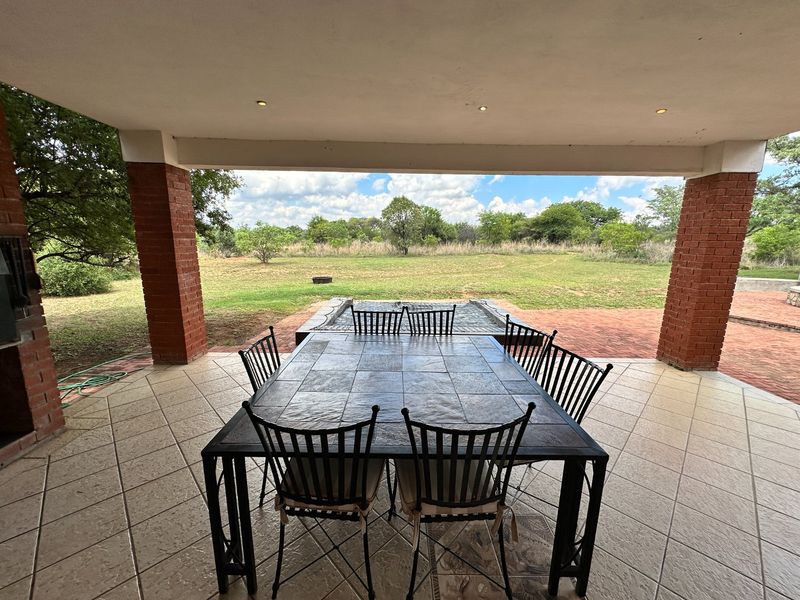 A spacious covered patio featuring tiled flooring, brick pillars, and a built-in braai, overlooking a large garden area.