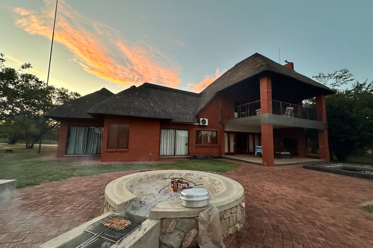 Rear view of a double-story brick house with a thatched roof, featuring a spacious paved patio area and a built-in circular fire pit.