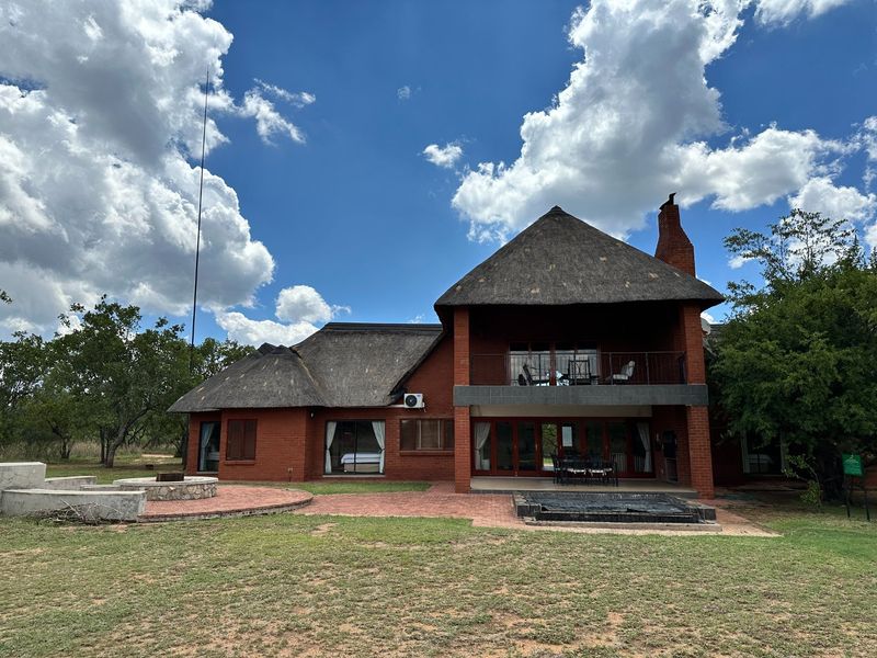 Rear view of a double-story brick house with a traditional thatch roof, featuring a balcony, ground-floor patio, and built-in outdoor braai area.