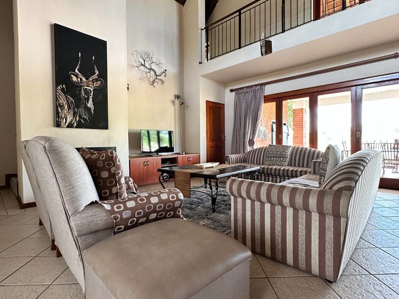 Spacious double-volume living room featuring a high thatched ceiling, mezzanine level with metal balustrade, tiled floors, and large sliding doors leading to the patio.