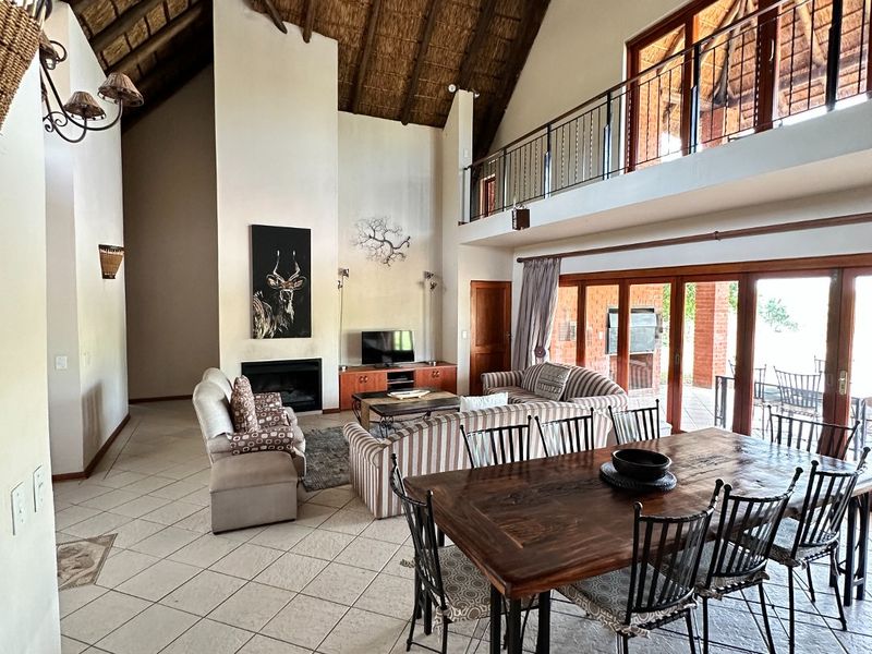 Spacious open-plan living and dining area featuring a dramatic vaulted thatched ceiling, mezzanine level, fireplace, and large sliding doors leading to the patio.