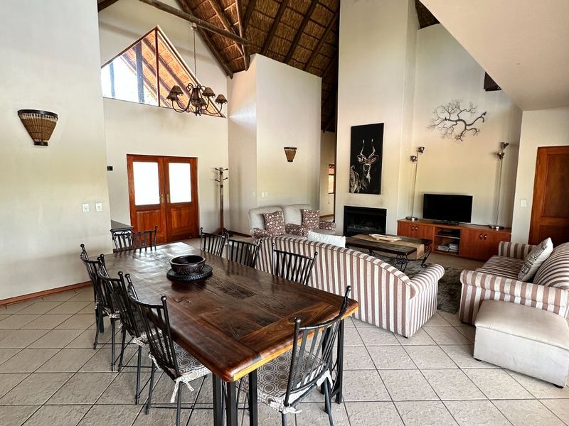 Spacious open-plan living and dining area featuring a high vaulted thatched ceiling with exposed timber trusses, tiled flooring, and a built-in fireplace.