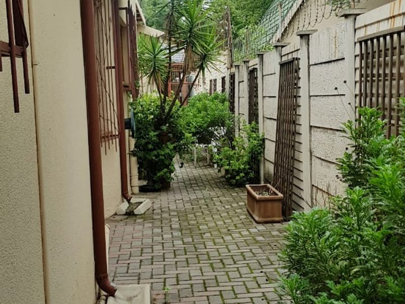 A narrow rear exterior walkway featuring paved flooring, perimeter boundary walls with security fencing, and functional drainage downpipes.