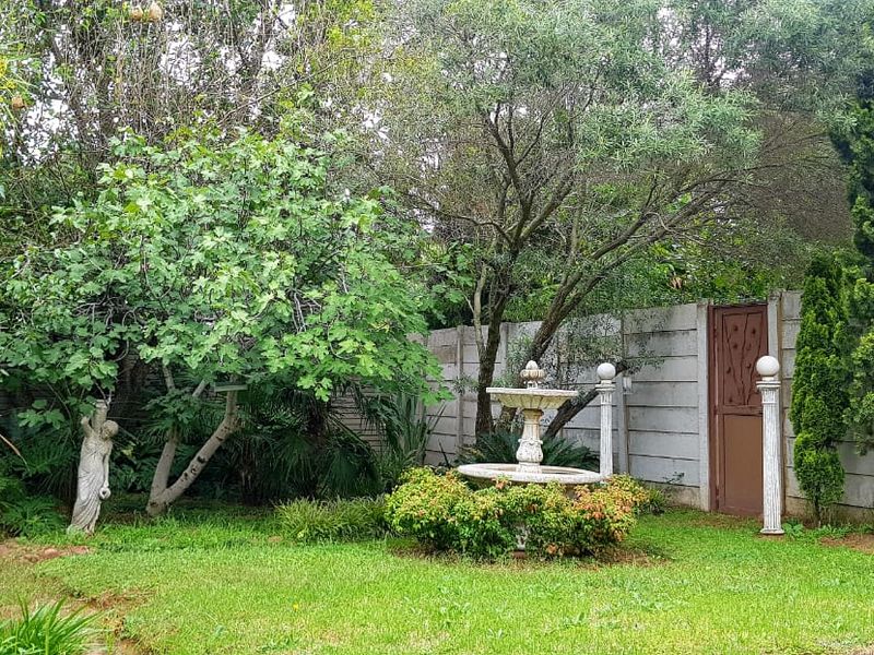 A lush, established garden featuring mature trees, a central stone fountain, and a secure precast concrete boundary wall with a pedestrian access gate.
