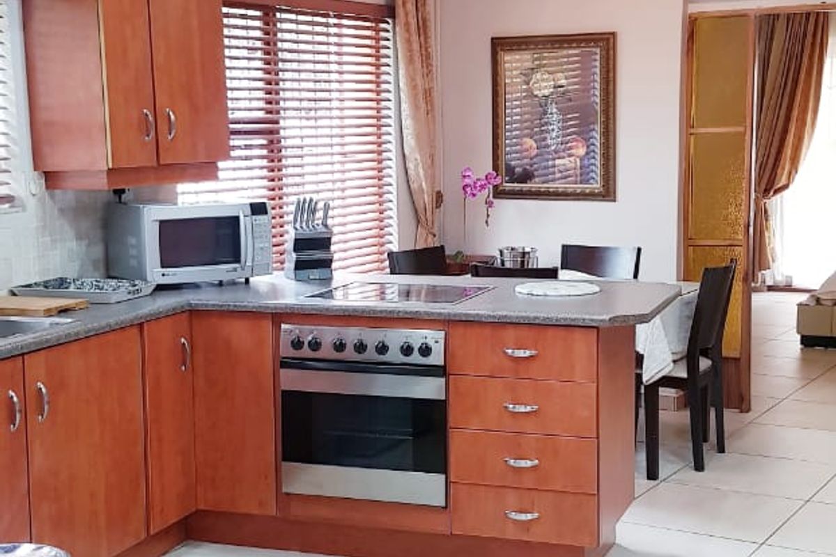 A functional kitchen featuring warm-toned wooden cabinetry, laminate countertops, built-in oven and hob, and neutral tiled flooring, leading into an adjacent dining area.