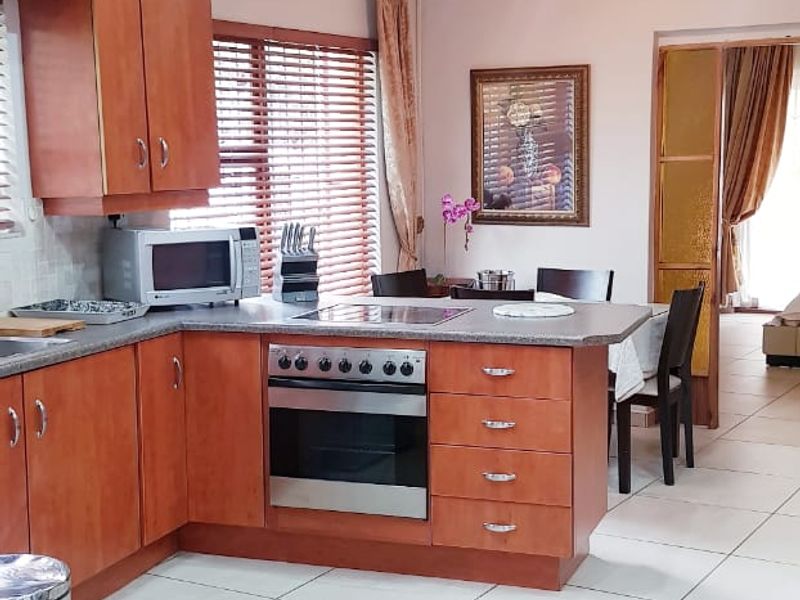 A functional kitchen featuring warm-toned wooden cabinetry, laminate countertops, built-in oven and hob, and neutral tiled flooring, leading into an adjacent dining area.