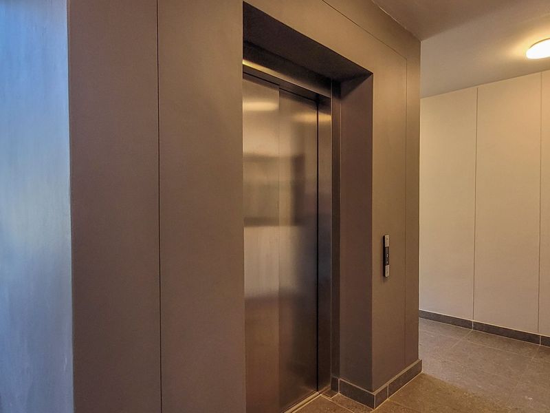 A modern, well-maintained apartment building hallway featuring a stainless steel elevator, tiled flooring, and an adjacent staircase with a handrail.
