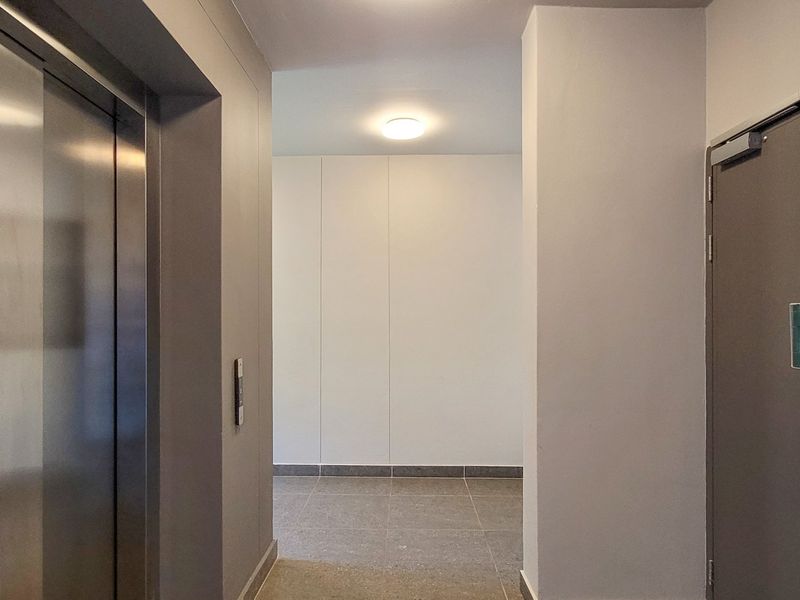 A clean and modern apartment building hallway featuring a stainless steel elevator, tiled flooring, and secure emergency access points.