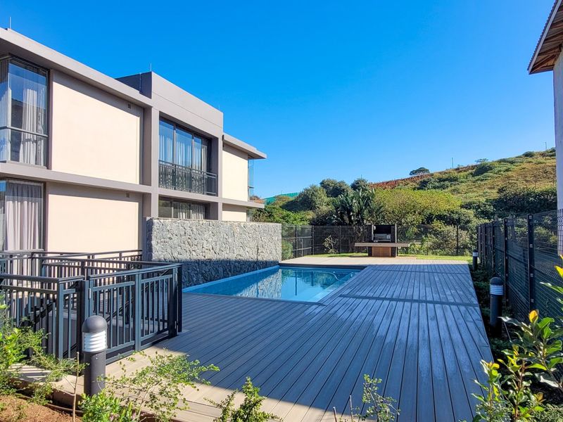 A well-maintained outdoor pool area featuring modern timber decking, a stone feature wall, and a built-in braai, set against a scenic hillside backdrop.