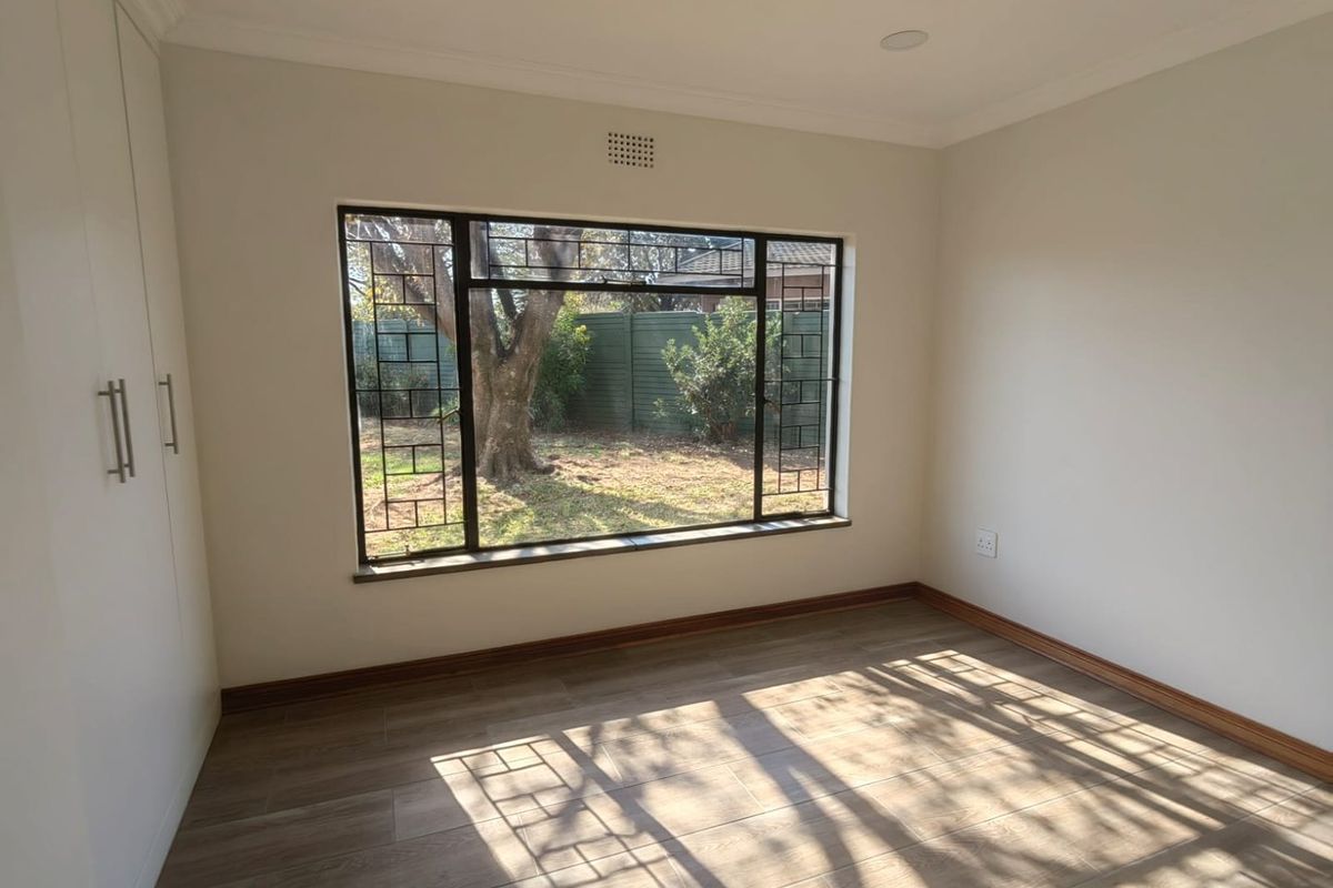 A bright, empty bedroom featuring modern wood-look tile flooring, built-in white cabinetry, and a large window with security bars overlooking the garden.